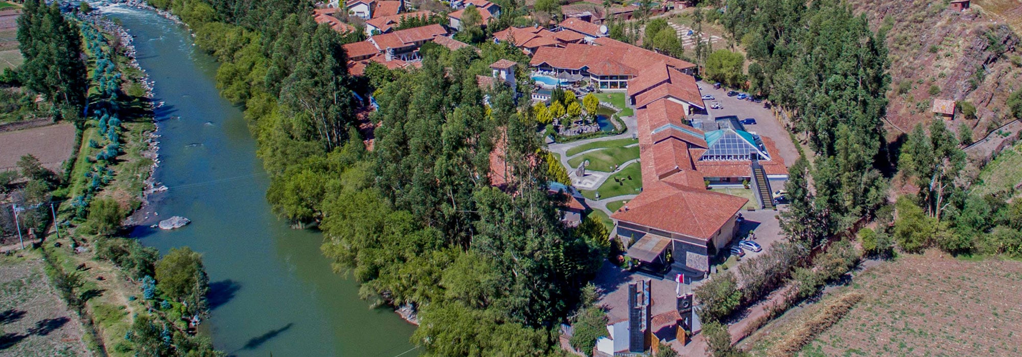 Aerial view of Cusco hotel complex with red-tiled roofs beside a river and lush vegetation | Ile Tours