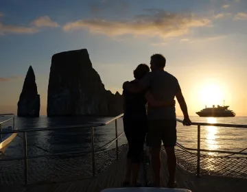 Couple standing on boat deck at sunset with Galapagos rock formations