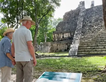 Senior couple walking along shaded route near Mayan ruins surrounded by trees and archaeological structures in Mexico | Ile Tours