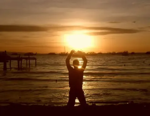 Silhouetted person stands on beach with arms raised toward golden sunset, boats and pier in background | Ile Tours