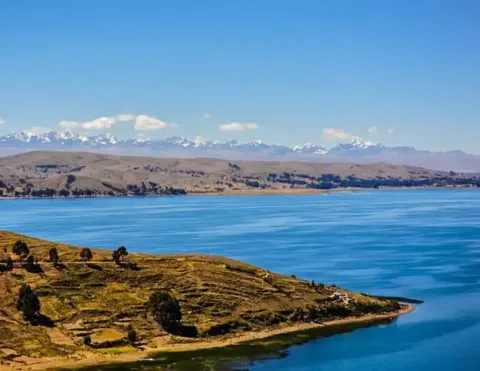 Blue waters of Lake Titicaca bordered by terraced land and trees, with snow-capped mountains in the distance | Ile Tours