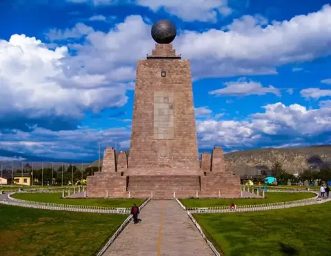 Stone monument with globe atop marks the equator near Quito, surrounded by visitors and mountain views | Ile Tours