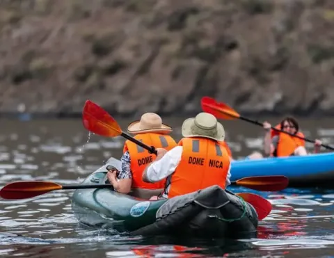 Group paddles inflatable kayaks wearing orange life vests, on calm water near rocky shoreline | Ile Tours