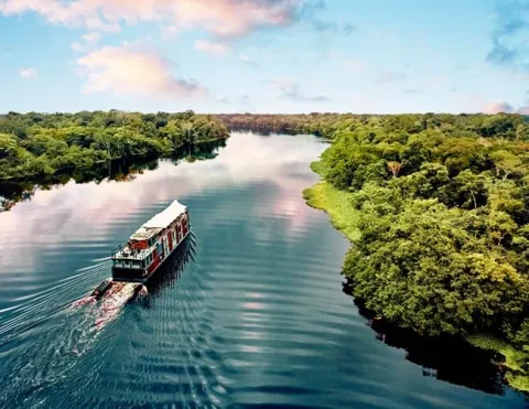 Barco turístico cruza río tranquilo rodeado de selva densa, bajo cielo rosado y reflejos dorados | Ile Tours