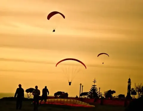 Paragliders soar through golden sunset skies above coastal cliffs, with silhouetted figures and lighthouse in scenic South American setting | Ile Tours