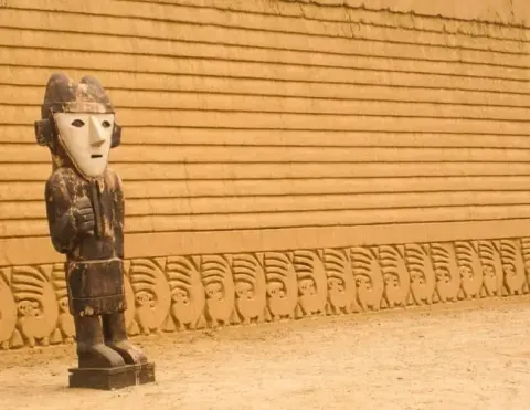 Wooden guardian statue stands before carved adobe wall with fish motifs at Chan Chan archaeological site in northern Peru | Ile Tours