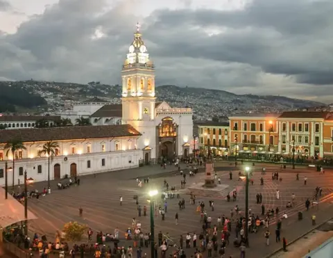 People gather in historic town square at dusk, with colonial church and bell tower lit against hilly cityscape | Ile Tours