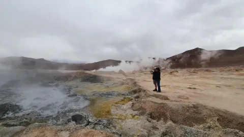 Explore Geothermal Wonders | Ile Tours A visitor observing steaming geothermal features in a rugged landscape with volcanic activity under a cloudy sky | Ile Tours
