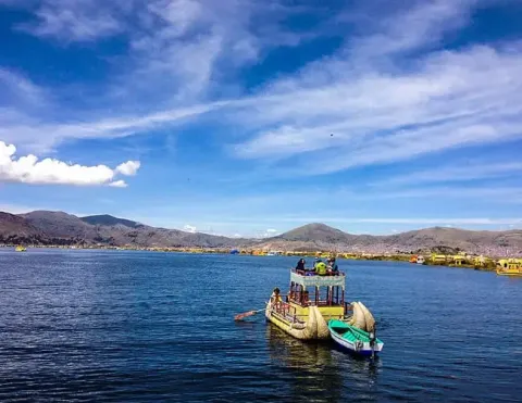 Embarcación tradicional de totora con toldo navega en el Lago Titicaca, con pasajeros a bordo y las coloridas islas Uros al fondo | Ile Tours
