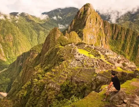 Explora la majestuosa Machu Picchu con Ile Tours Viajero sentado admirando la impresionante vista de Machu Picchu, con montañas verdes exuberantes de fondo | Ile Tours