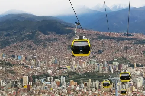 Discover the Unique Cable Car System of La Paz | Ile Tours A panoramic view of a city with yellow cable cars traveling above it, showcasing a mix of urban and mountainous landscapes. | Ile Tours