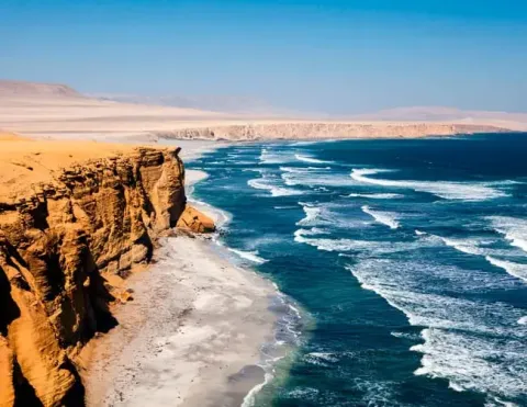 Acantilados rojizos y desierto se encuentran con el océano azul profundo en la costa peruana, con olas paralelas y cielo despejado | Ile Tours