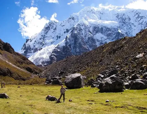Hiker exploring a grassy valley with towering snow-capped mountains in the background under a clear blue sky | Ile Tours