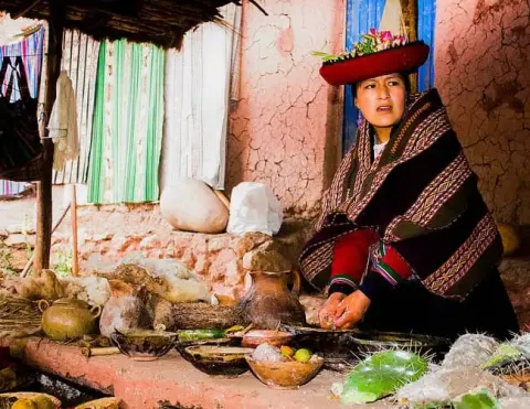 Mujer andina con vestimenta tradicional prepara elementos naturales en mercado rústico al aire libre, con muros de adobe y textiles culturales en Perú | Ile Tours