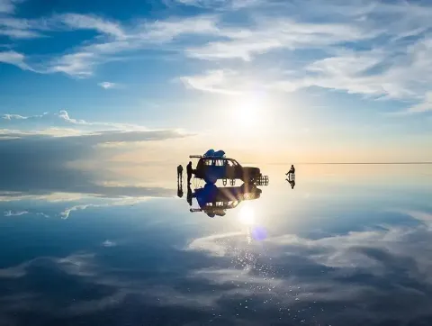 Explore Breathtaking Natural Wonders with Ile Tours Reflection of a vintage car and people on a vast salt flat at sunset, with a stunning sky and sun illuminating the scene | Ile Tours