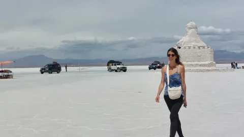 Uma viajante caminhando pelas extensas salinas do Salar de Uyuni, com veículos ao fundo e as icônicas esculturas de sal, mostrando a paisagem surrealista da Bolívia | Ile Tours