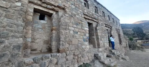 A stone building with windows at an archaeological site, featuring a person standing beside it, capturing the essence of ancient architecture in the Andean region of Bolivia | Ile Tours
