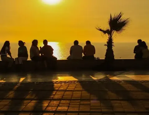 Un sereno atardecer sobre el océano, con siluetas de personas sentadas juntas a lo largo de un Paseo costero, palmeras enmarcando la escena, creando un ambiente cálido y acogedor | Ile Tours
