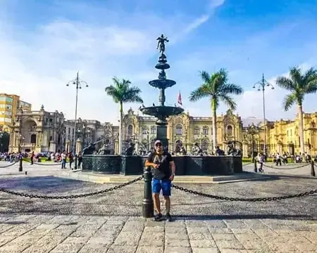 Una persona posando frente a una gran fuente en una plaza animada, rodeada de palmeras, con edificios históricos al fondo y un cielo azul claro | Ile Tours