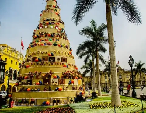 Una vibrante torre decorativa hecha de coloridos artesanías y objetos tradicionales, situada en una plaza verde adornada con palmeras y arreglos florales, mostrando la riqueza cultural de Lima | Ile Tours