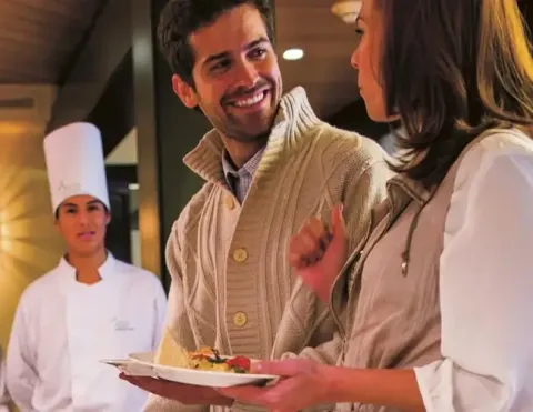 A smiling man holding a plate of food while engaging in conversation with a woman, with a chef in the background | Ile Tours