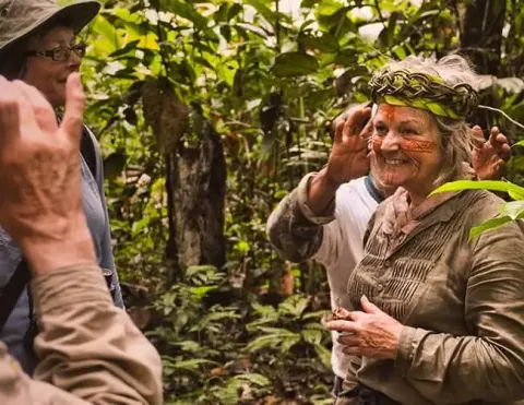 Un grupo de personas interactuando en un entorno de bosque exuberante, con una mujer usando una corona tradicional y decorada con pintura facial tribal, sonriendo. | Interacción Cultural