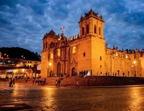 The Cusco Cathedral illuminated at night in the Plaza de Armas, with people walking around and a dramatic sky.