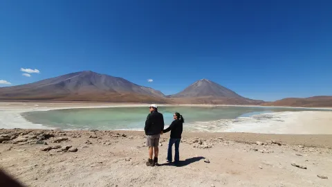 A couple standing by the stunning green lagoon in the Altiplano region, with majestic volcanoes in the background under a clear blue sky, capturing the beauty of Bolivia's natural landscapes | Ile Tours