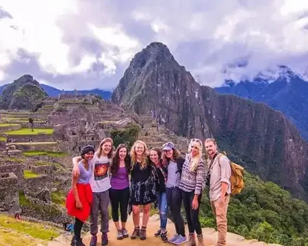 A group of eight travelers posing together with the stunning backdrop of Machu Picchu, showcasing the ancient ruins and surrounding mountains on a cloudy day | Ile Tours