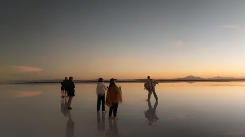 A group of people standing on a reflective salt flat at sunset, with soft colors in the sky and mountains in the background, creating a serene and captivating atmosphere. | Ile Tours