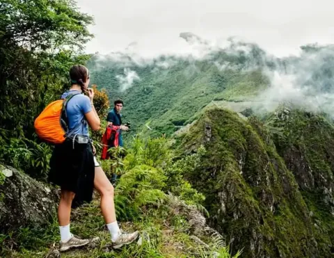 Two hikers exploring the stunning landscape of Machu Picchu, one taking a photo while the other gazes at the breathtaking mountains and clouds surrounding the area | Ile Tours