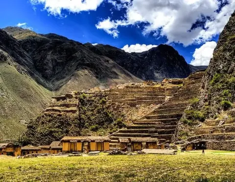 Vista cênica das ruínas incas de Ollantaytambo, com campos em terraços e antigas estruturas de pedra contra uma paisagem montanhosa e um céu azul com nuvens | Ile Tours