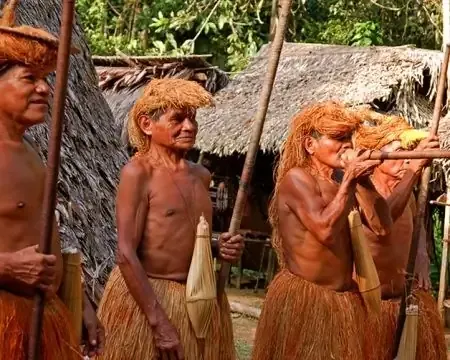 Un grupo de hombres indígenas vistiendo atuendos tradicionales hechos de materiales naturales, de pie con lanzas y flautas, rodeados de una aldea boscosa. | Culturas Indígenas