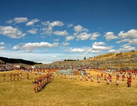 Uma foto ampla do festival Inti Raymi em Sacsayhuaman, Cusco, com artistas no campo e uma grande multidão assistindo.