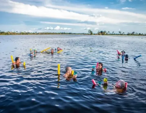 Un grupo de personas disfrutando de un día divertido nadando en un río sereno, usando fideos de piscina, bajo un cielo azul brillante | Ile Tours