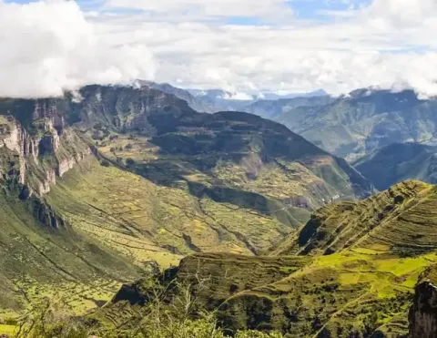 Una vista impresionante de valles verdes y montañas empinadas, con parches de tierras cultivadas y nubes esponjosas flotando sobre el paisaje andino, mostrando la belleza del paisaje natural de Perú | Ile Tours