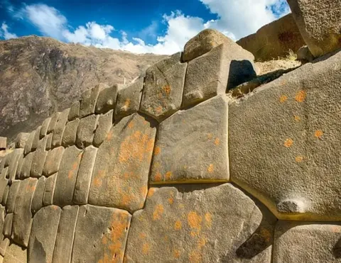 Uma vista de perto de uma antiga parede de pedra inca, mostrando o trabalho de artesanato preciso e os padrões únicos das pedras, com um fundo das montanhas dos Andes e um céu azul | Ile Tours