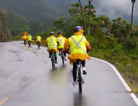 Um grupo de ciclistas usando roupas de chuva amarelas brilhantes pedalando por uma estrada molhada nas montanhas, cercados por uma vegetação exuberante e névoa, mostrando a aventura do ciclismo em diversas condições climáticas no Peru | Ile Tours