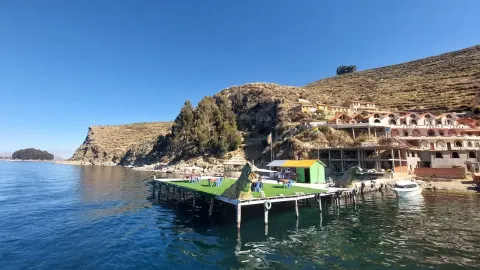 A picturesque view of a dock with green grass and small boats, surrounded by stunning hills and charming buildings along the water's edge, under a clear blue sky. | Ile Tours