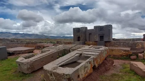 Ancient stone structures at a historical site, surrounded by grassy fields and dramatic clouds in the sky, showcasing remnants of an ancient civilization. | Ile Tours