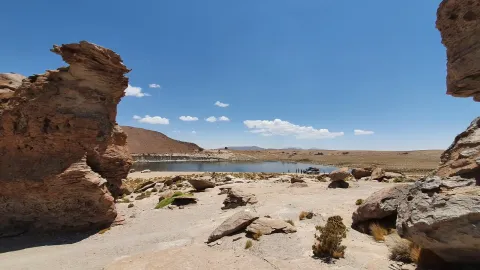 A scenic view of rock formations and a tranquil lagoon in the Altiplano region of Bolivia, with a clear blue sky and distant mountains, showcasing the unique natural beauty of the area | Ile Tours