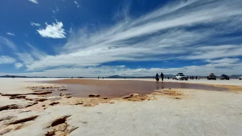 Una vista amplia del Salar de Uyuni con una piscina salina poco profunda rodeada de visitantes y vehículos bajo un cielo dramático | Ile Tours