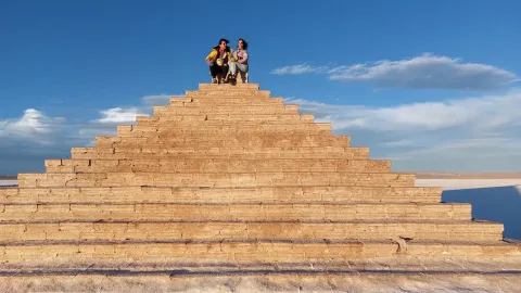Dos personas sentadas en la cima de una formación en forma de pirámide hecha de bloques de sal en el Salar de Uyuni, frente a un cielo claro con nubes suaves | Ile Tours