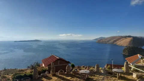 A scenic view of a tranquil lake surrounded by mountains, with charming houses and stone tables in the foreground, bathed in warm sunlight under a clear blue sky. | Ile Tours