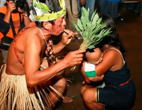 Un chamán realizando un ritual mientras sostiene a un bebé, rodeado de vestimenta tradicional y vegetación.