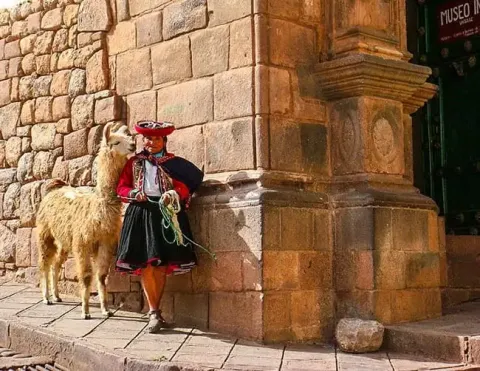 A person in traditional Peruvian attire standing beside a llama near a stone wall, showcasing the vibrant cultural heritage.