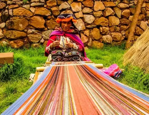 Uma mulher vestindo roupas tradicionais tecendo tecidos coloridos em um ambiente rural, com paredes de pedra e vegetação ao fundo | Ile Tours