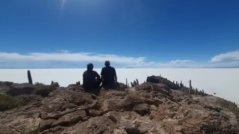 Dos personas sentadas en un afloramiento rocoso con vistas a las extensas salinas blancas del Salar de Uyuni, con cactus en el primer plano y un cielo azul brillante | Ile Tours